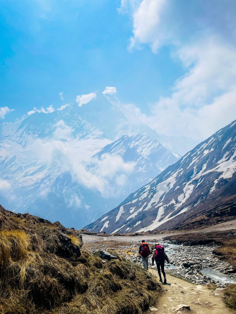 Two backpackers trekking on a dirt trail toward a snow-capped Himalayan mountain.