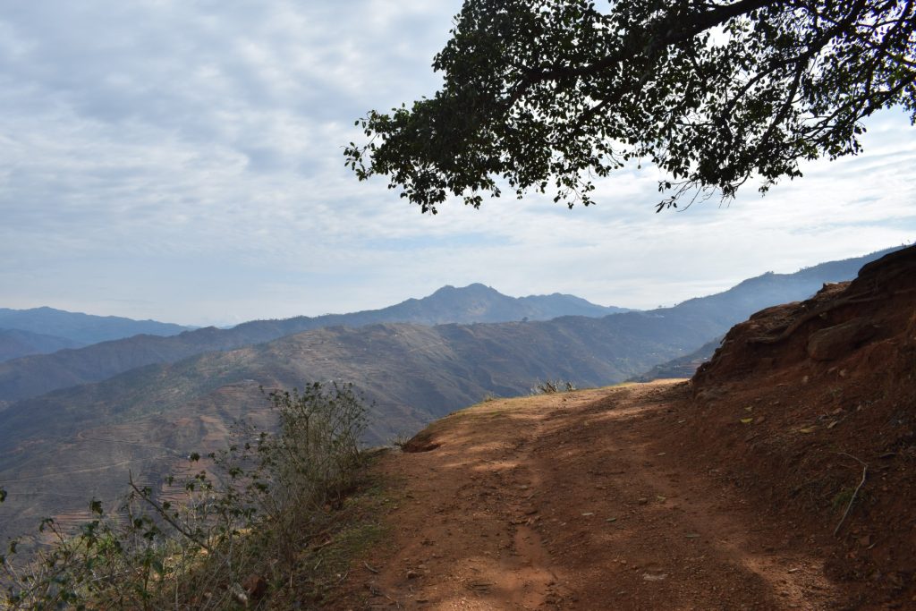  "A narrow dirt hiking trail winding along a rugged mountain ridge overlooking the vast, hazy valley and peaks of Limi Valley in the Humla district of Nepal.