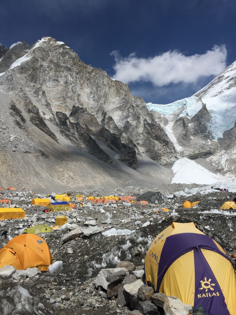 Yellow and purple mountaineering tents set up at Everest Base Camp with rocky terrain, snow, and steep mountain peaks under a blue sky.