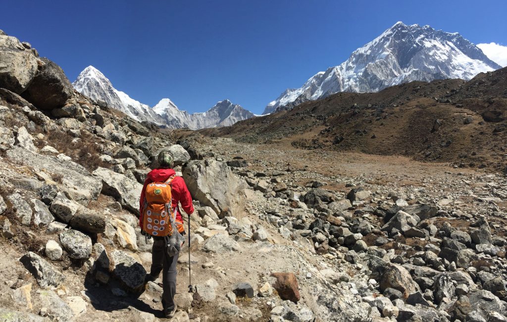 Hiker with a red jacket trekking on a rocky trail toward Mount Everest under a clear blue sky.