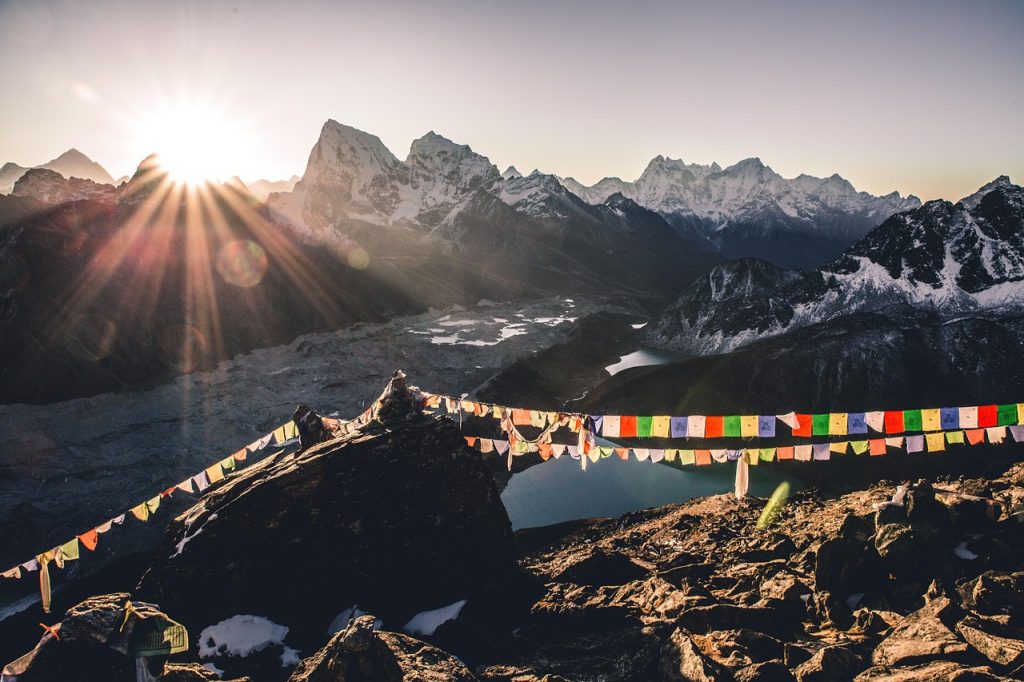 A breathtaking sunrise over the snow-capped Himalayan mountains, viewed from the rocky summit of Gokyo Ri, featuring vibrant Buddhist prayer flags strung across the foreground and a glacier-fed lake valley below.