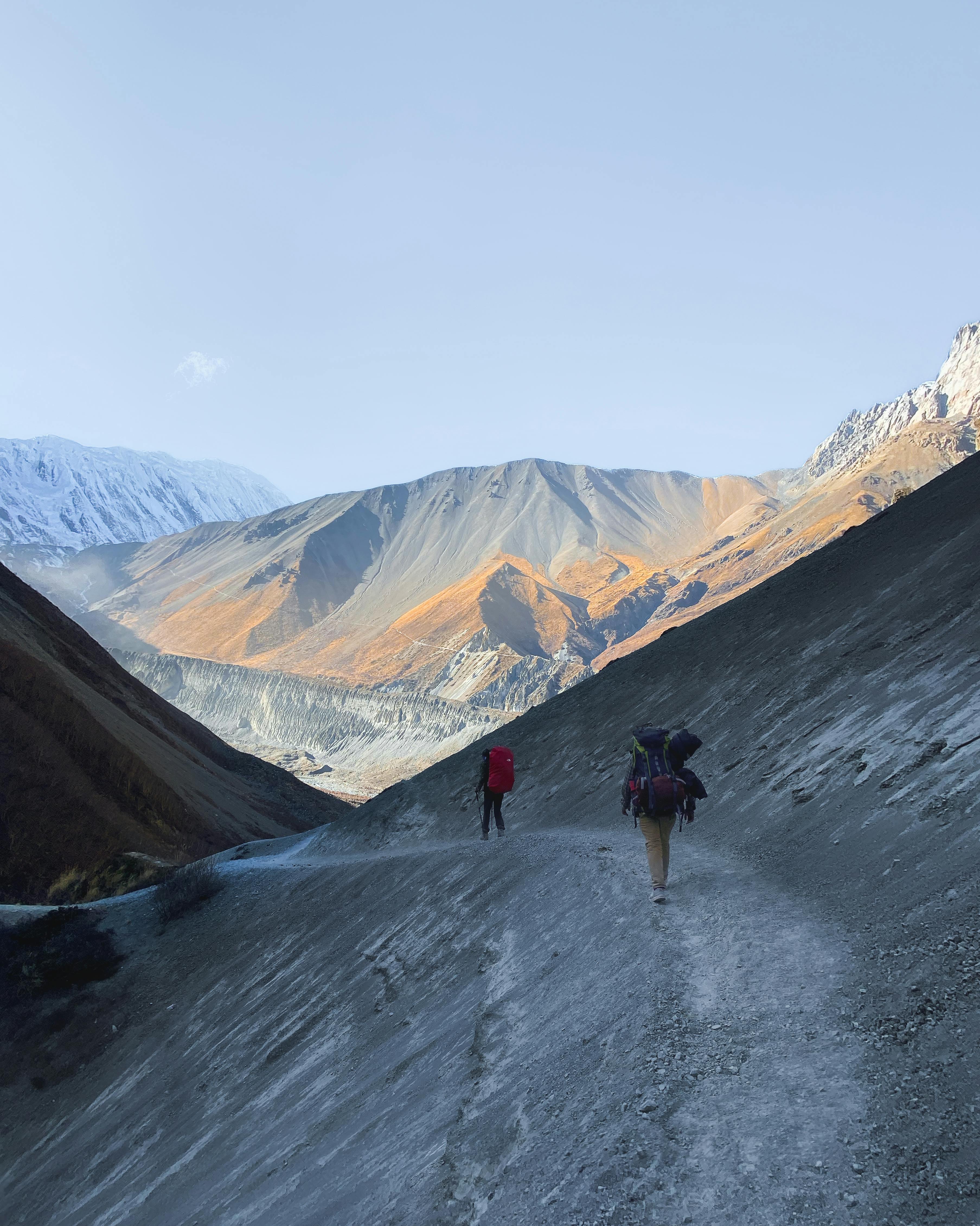 Two hikers trekking on a rugged mountain path toward Tilicho Lake in the Annapurna region of Nepal.