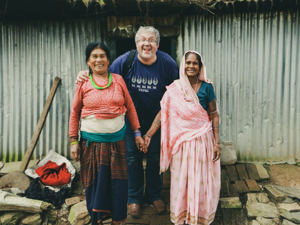 Man with two Nepali women in traditional attire standing in front of a corrugated metal house in Nepal.