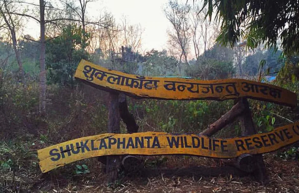  A rustic, two-tiered yellow wooden sign with hand-painted black lettering in Nepali and English reading "Shuklaphanta Wildlife Reserve," set against a backdrop of lush green trees and natural forest vegetation in Nepal.