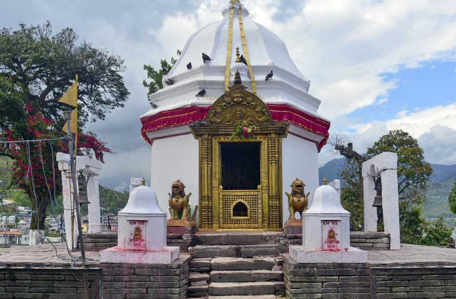 A white octagonal Shikhara-style Hindu temple dome, known as the Bindhyabasini Temple in Pokhara, Nepal, featuring a gilded golden doorway entrance guarded by two small stone lion statues on a stone platform under an overcast sky.