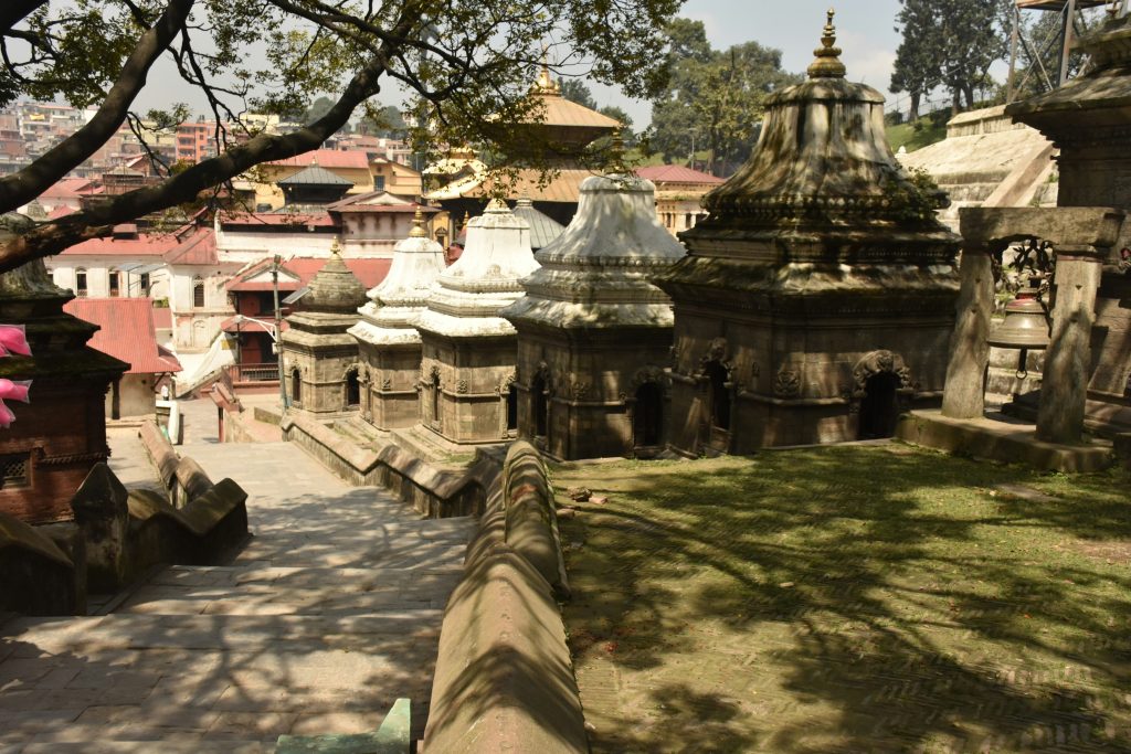 A stone stairway descends toward a row of historical stone shrines at the Pashupatinath Temple complex in Kathmandu, Nepal, shaded by tree branches on a sunny day.