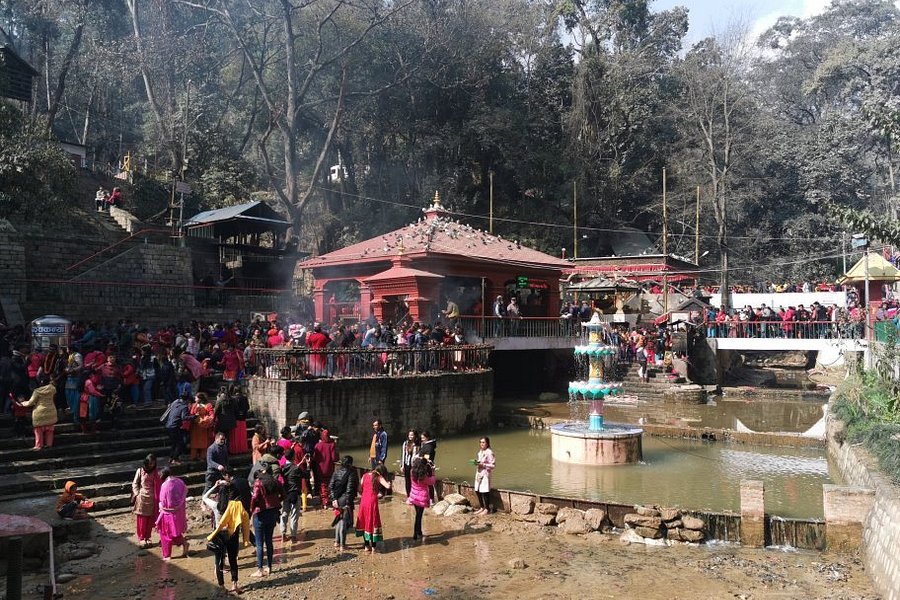 Crowded Hindu devotees gathering at the sacred Dakshinkali Temple in Pharping, Nepal, featuring the distinctive red-roofed main shrine and central water fountain.
