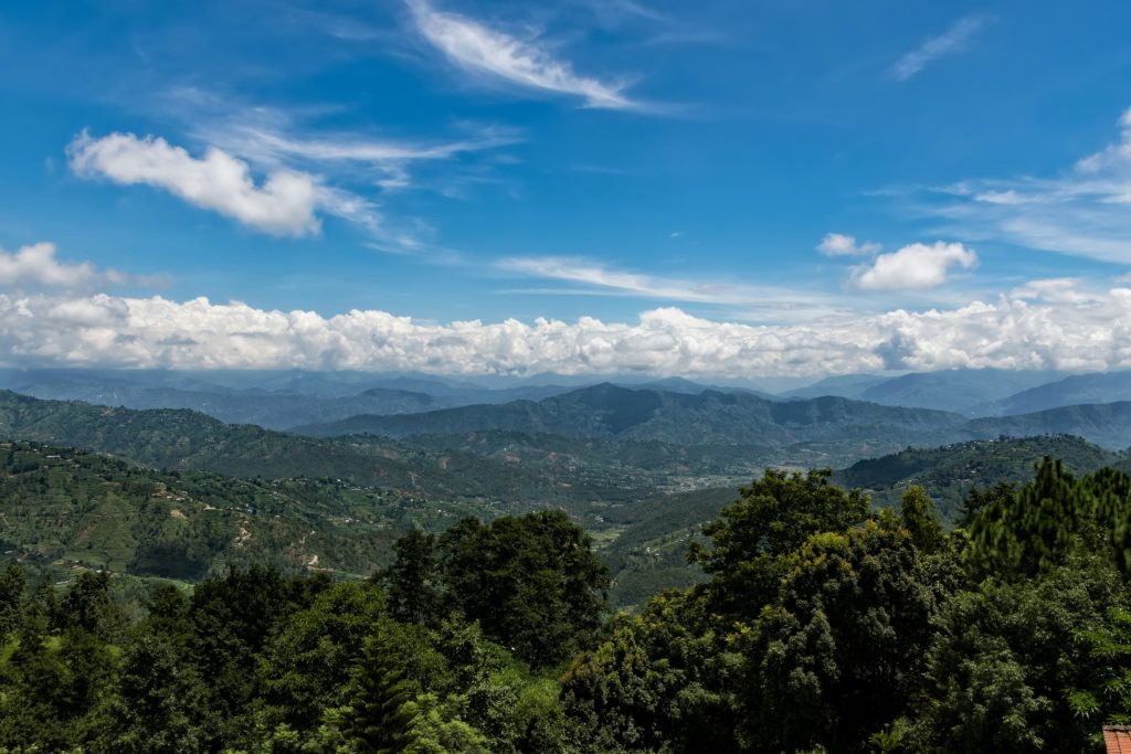 Lush green terraced hills of Dhulikhel, Nepal, overlooking a deep valley with the Himalayan foothills under a bright blue sky with scattered clouds.