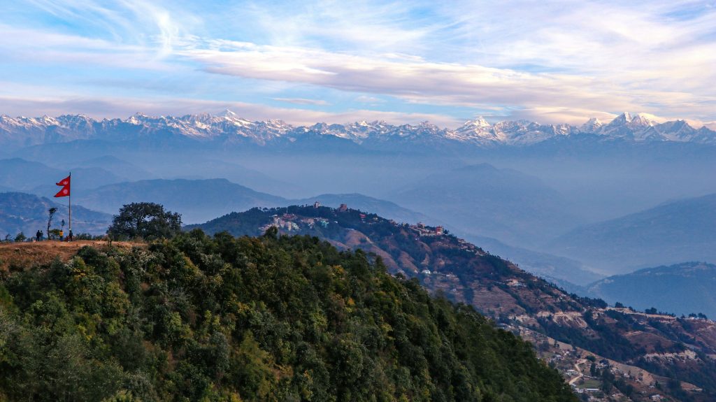 Wide panoramic view of the snow-capped Himalayan mountain range under a blue sky from Nagarkot, Nepal, featuring a Nepalese flag in the foreground.