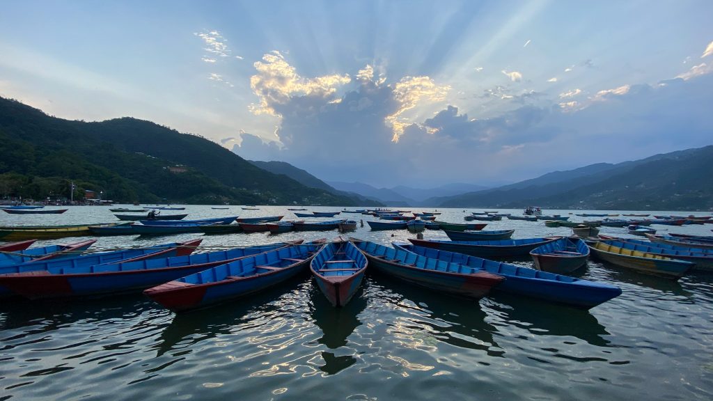 Colorful traditional wooden boats moored on the calm waters of Phewa Lake in Pokhara, Nepal, under a dramatic sunset sky with light rays breaking through the clouds.