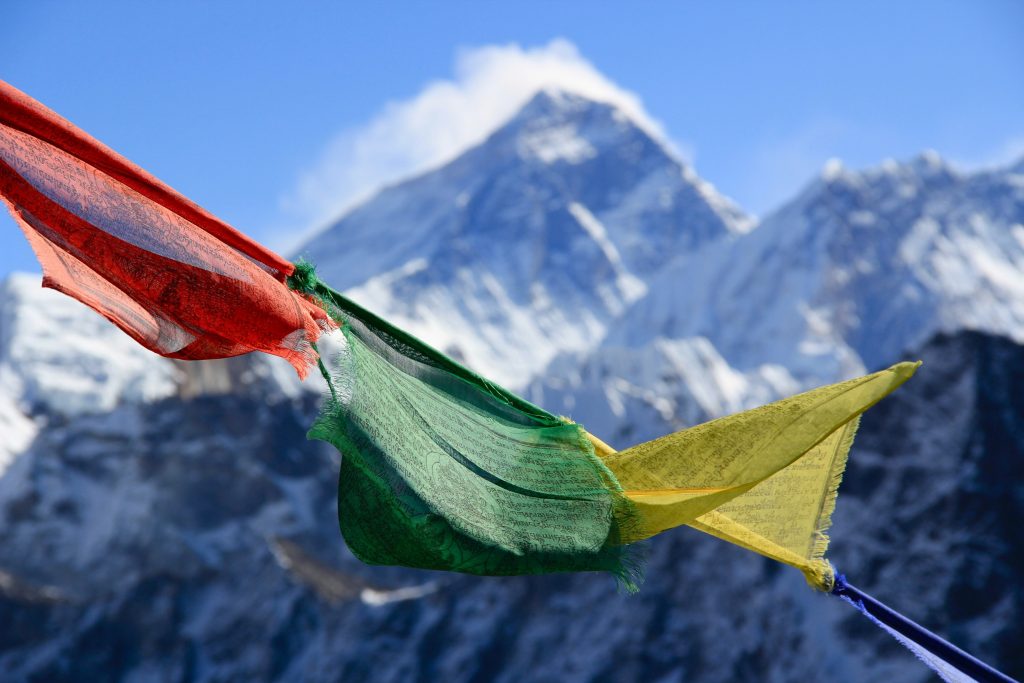  Vibrant Buddhist prayer flags fluttering on a rocky ridge with snow-capped Himalayan mountain peaks and a high-altitude Sherpa village in the background.