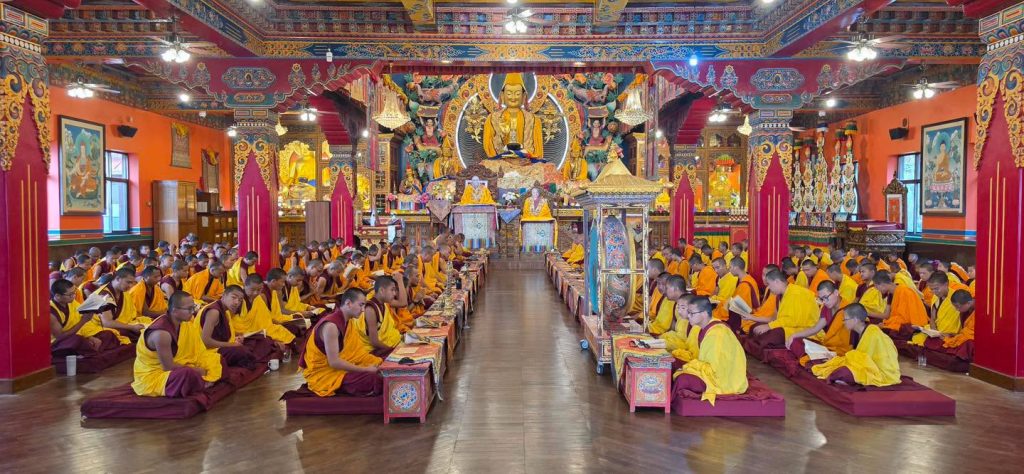 Tibetan Buddhist monks in maroon and yellow robes sit in rows participating in a morning puja prayer ceremony inside the intricately decorated main prayer hall of Kopan Monastery, Kathmandu, Nepal, with a large golden Buddha statue in the background.