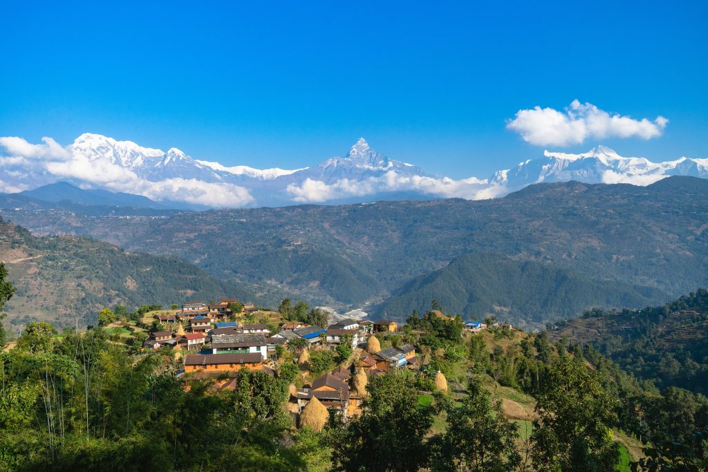 A scenic view of a traditional hillside village in Nepal, with the snow-covered peaks of the Annapurna mountain range and Machhapuchhare (Fishtail) rising under a clear blue sky.