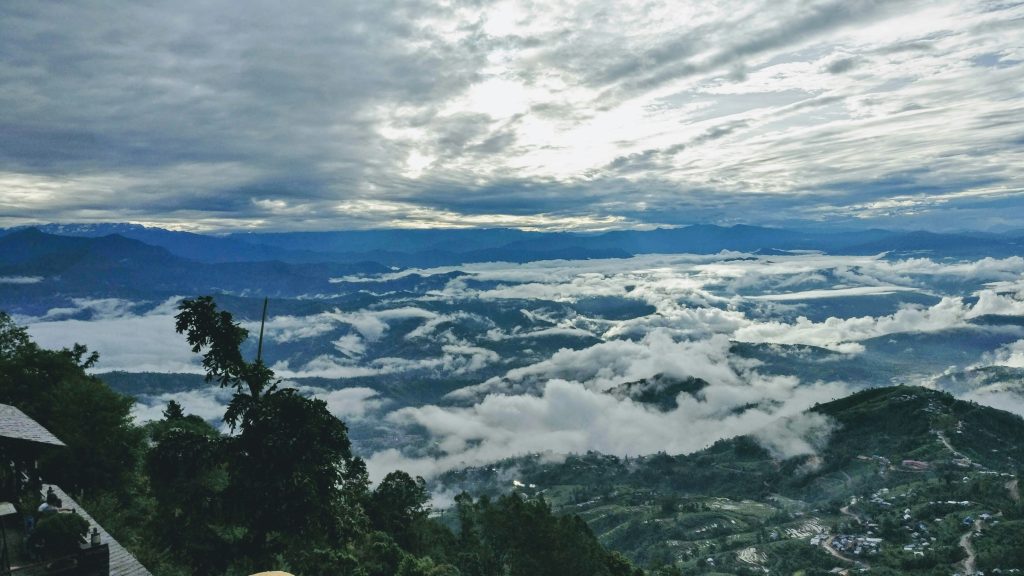 Panoramic view of the Himalayas from Nagarkot, Nepal, showing a dramatic sea of clouds over rolling green hills under a cloudy sky.