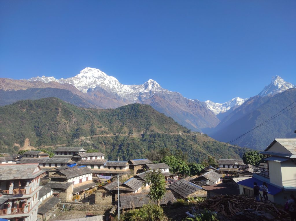 Traditional stone houses of Ghandruk village in Nepal with a panoramic view of Annapurna South and Machhapuchhre (Fishtail) peaks under a clear blue sky.
