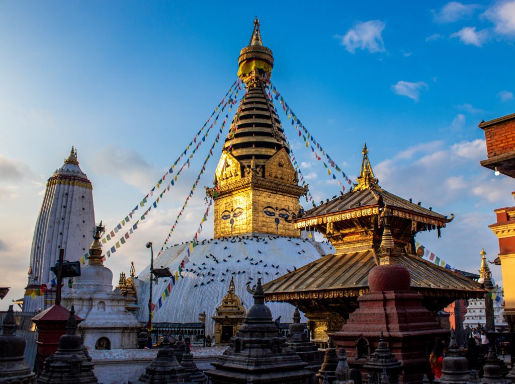  View of the golden Swayambhunath Stupa spire and white dome decorated with Buddha eyes, surrounded by colorful prayer flags and smaller shrines on a hilltop in Kathmandu.