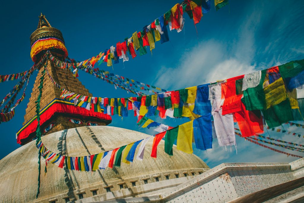 Massive white dome of Boudhanath Stupa in Kathmandu under a blue sky, draped with vibrant colorful Tibetan prayer flags.