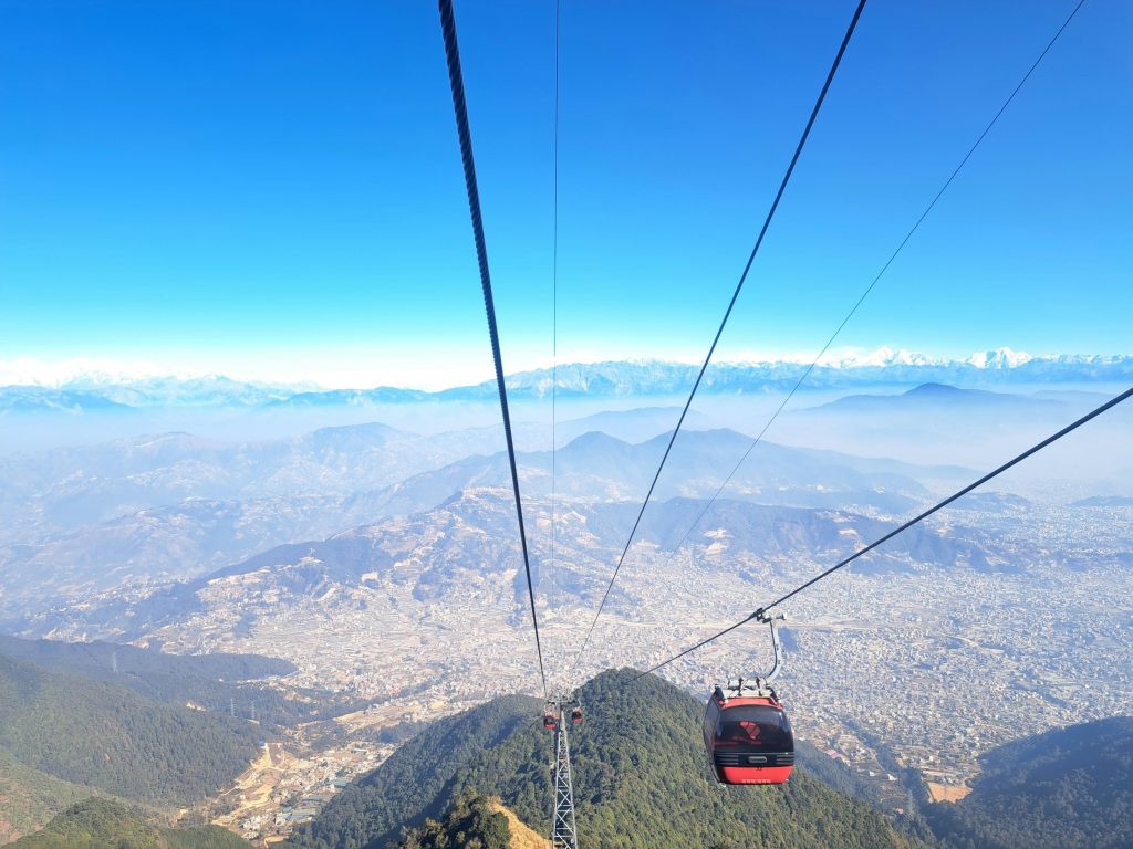 Red cable car gondola ascending Chandragiri Hills with a panoramic view of Kathmandu Valley and the snow-capped Himalayan mountain range under a clear blue sky.