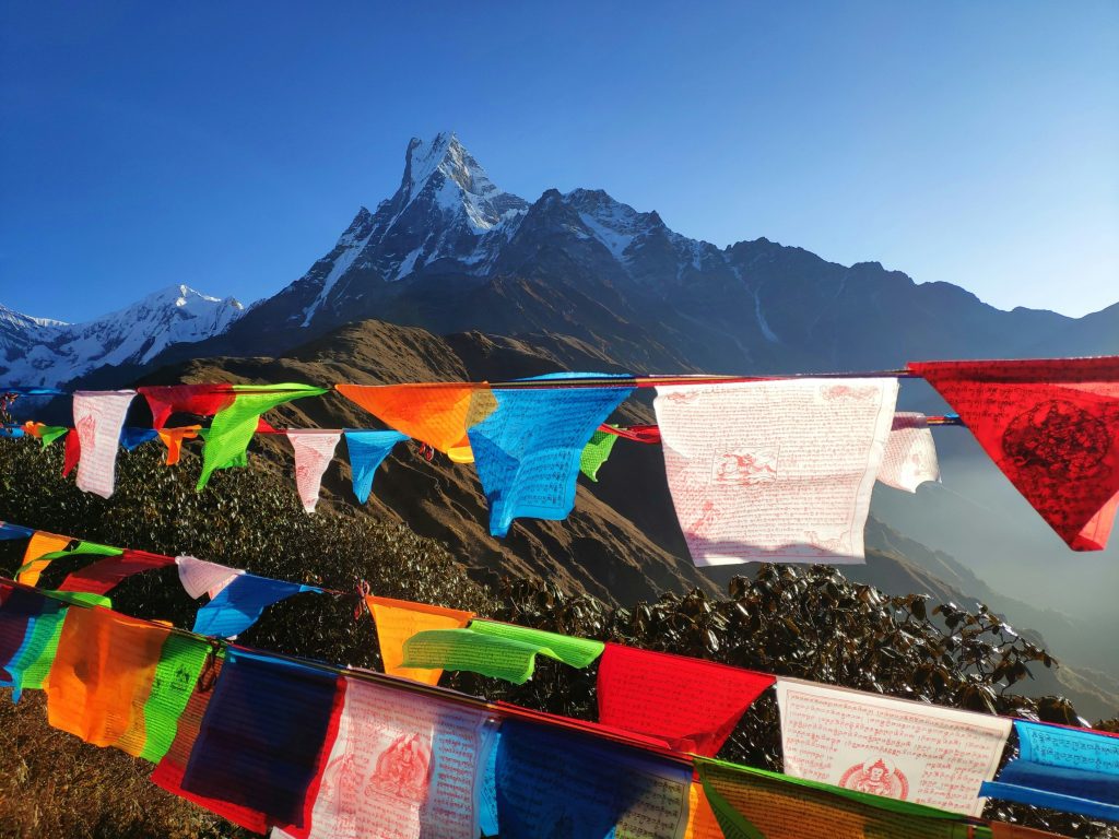 Vibrant Buddhist prayer flags fluttering in the foreground with the sacred Machhapuchhre (Fishtail Mountain) peak in the Annapurna Himalayas of Nepal under a clear blue sky.