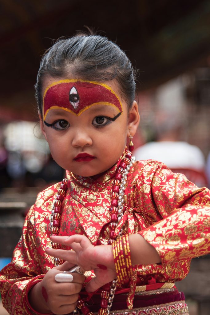  A young girl dressed as the Kumari living goddess in Nepal, wearing traditional red and gold silk attire, a painted third eye on her forehead, and intricate eye makeup.