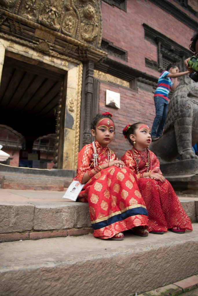 Two young Nepalese girls dressed in traditional red and gold silk attire and sacred face paint, participating in a Kumari ritual at a historic temple in Kathmandu.