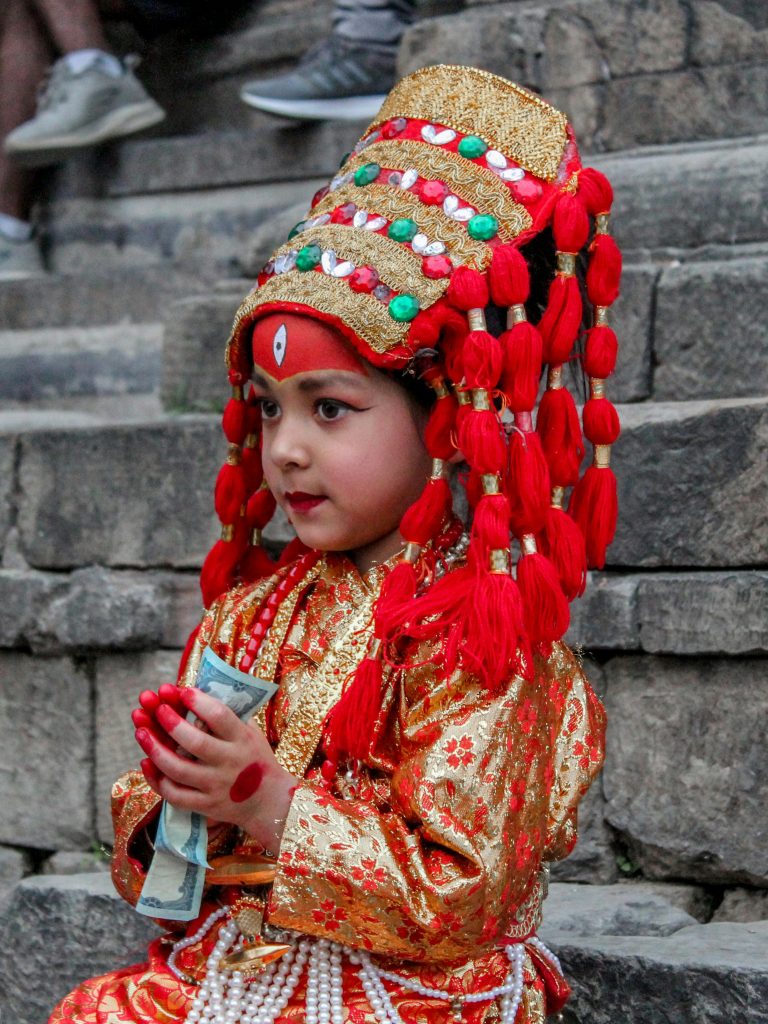 A young child dressed as a Kumari (Living Goddess) in traditional red and gold Nepali attire, wearing an ornate headdress with red tassels and holding money, seated on stone steps.