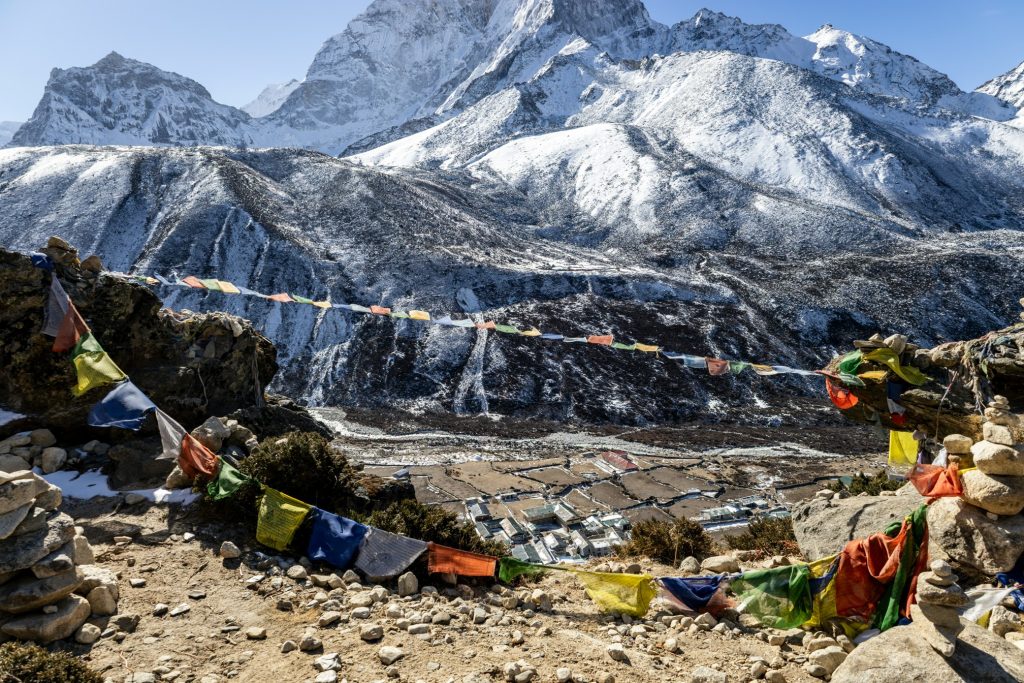Vibrant Buddhist prayer flags fluttering on a rocky ridge with snow-capped Himalayan mountain peaks and a high-altitude Sherpa village in the background.