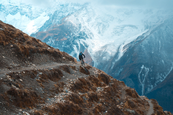 The Ultimate Adventure Guide In Nepal: Cyclist riding a mountain bike on a narrow cliffside dirt trail in the snow-capped Himalayan mountains