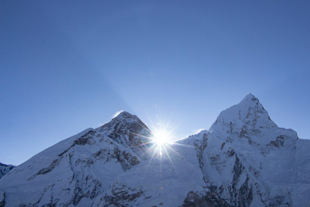 Sunburst rising over snow-covered Himalayan mountain peaks against a clear blue sky